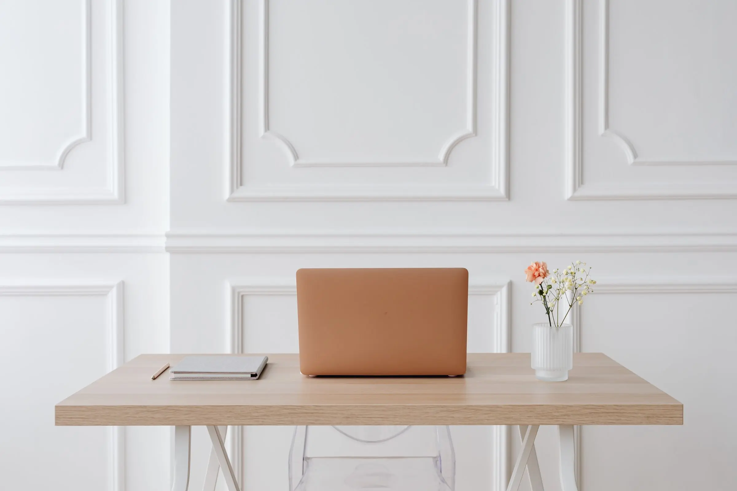 A tidy wooden desk with a laptop, a notebook and a small vase of flowers against a white panelled wall
